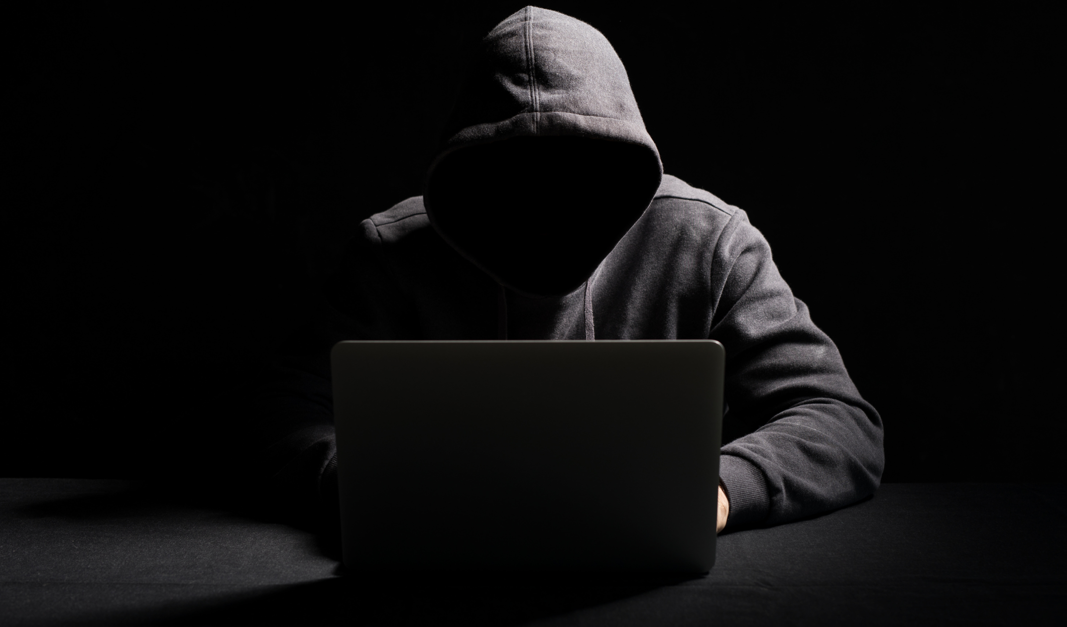 Hooded figure in shadows in front of a laptop to represent ghost students identity theft schemes.