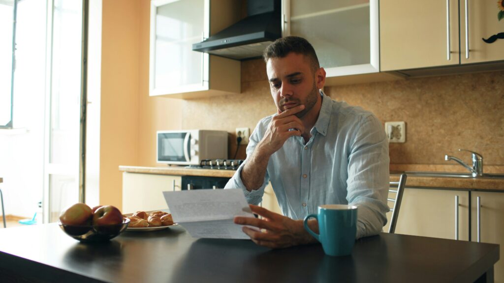 Man in a light blue shirt sits at a kitchen table reading a document, with a coffee mug and plate of croissants nearby while sunlight streams in from a window.